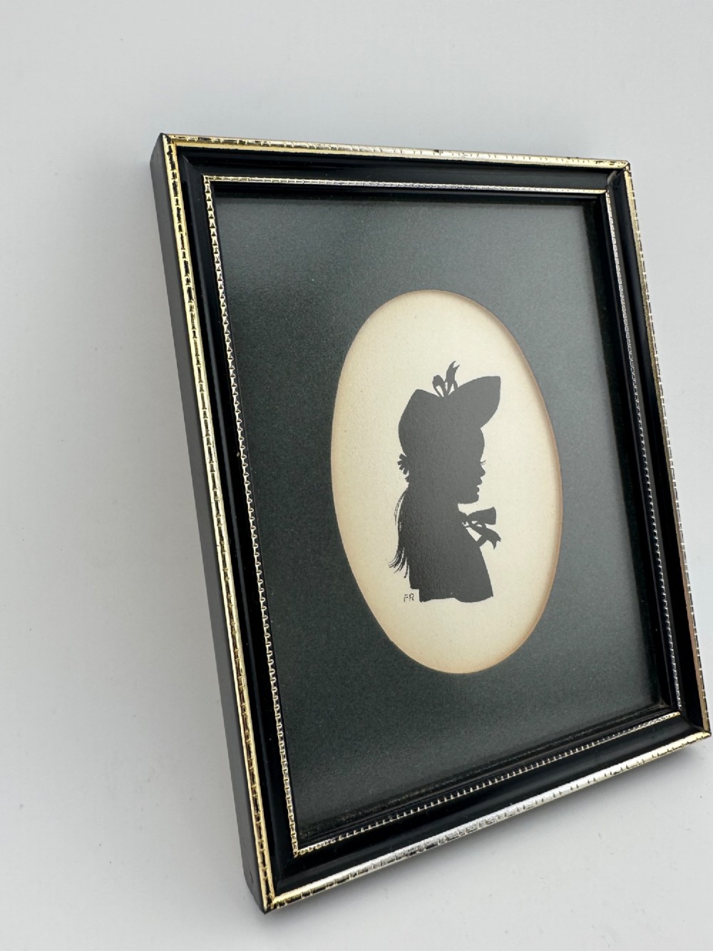 A framed Art Deco silhouette portrait depicting a lady in profile with a decorative hat inside an oval cream mount surrounded by a black and gold frame. View III