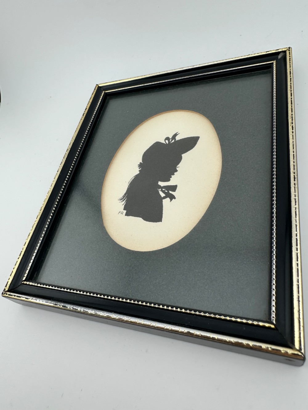 A framed Art Deco silhouette portrait depicting a lady in profile with a decorative hat inside an oval cream mount surrounded by a black and gold frame. View VII