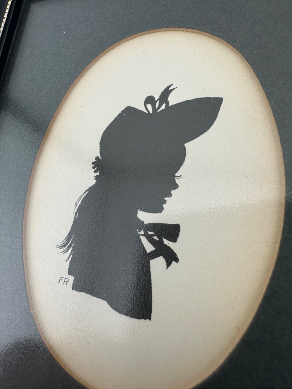 A framed Art Deco silhouette portrait depicting a lady in profile with a decorative hat inside an oval cream mount surrounded by a black and gold frame. View VIII