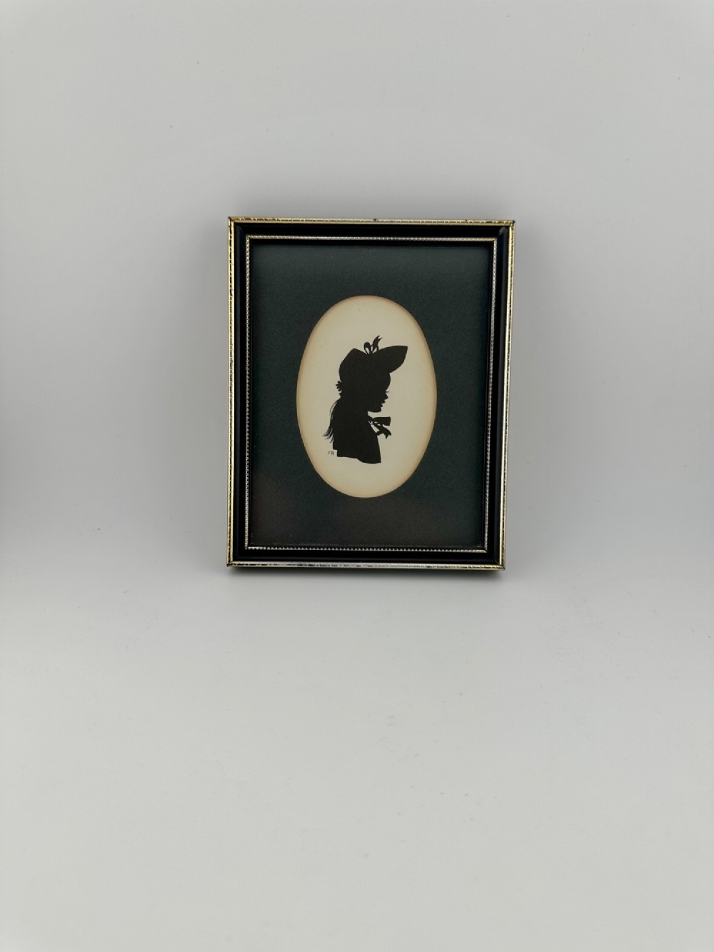 A framed Art Deco silhouette portrait depicting a lady in profile with a decorative hat inside an oval cream mount surrounded by a black and gold frame. View X