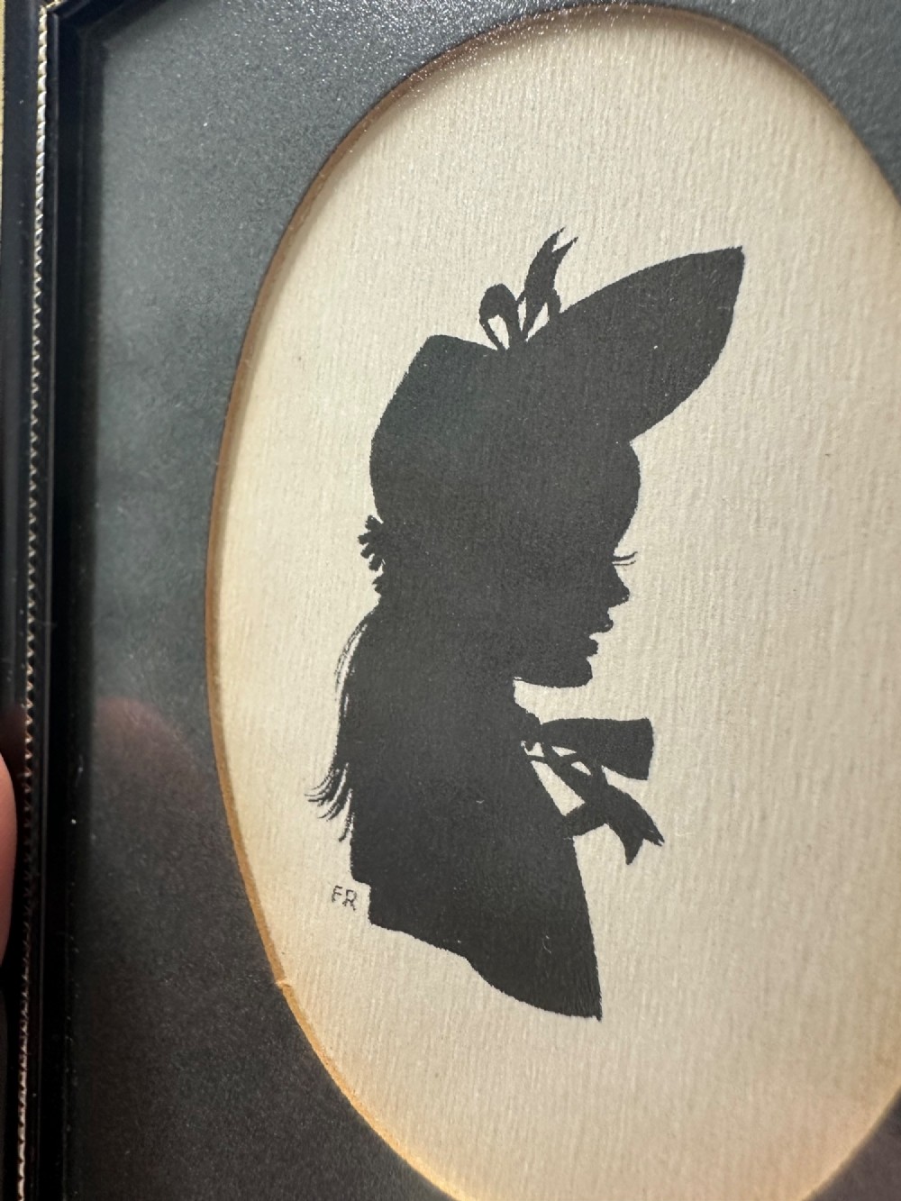 A framed Art Deco silhouette portrait depicting a lady in profile with a decorative hat inside an oval cream mount surrounded by a black and gold frame. View XI