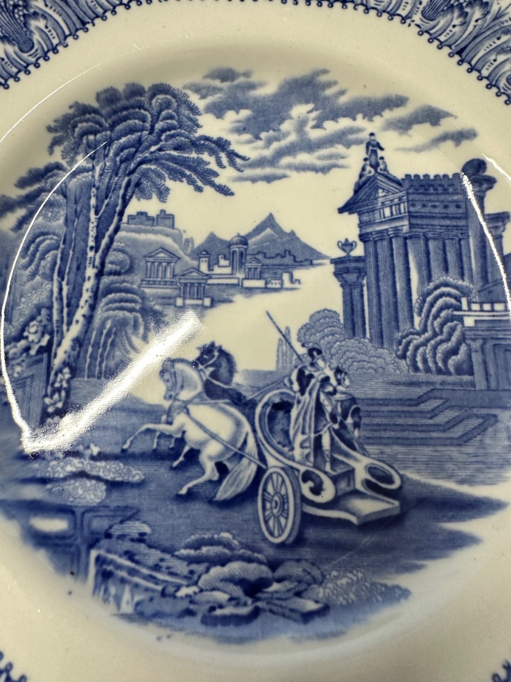 An octagonal-edged blue and white transferware plate featuring a central classical chariot scene surrounded by an ornate feather-patterned border. View X