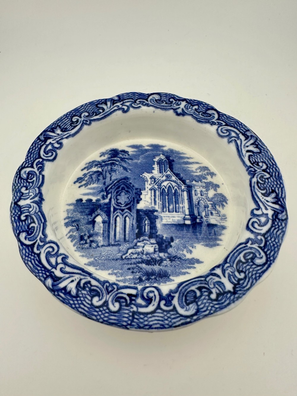 A circular blue and white transferware ceramic dish featuring a central printed depiction of abbey ruins framed by a decorative scrolled border.