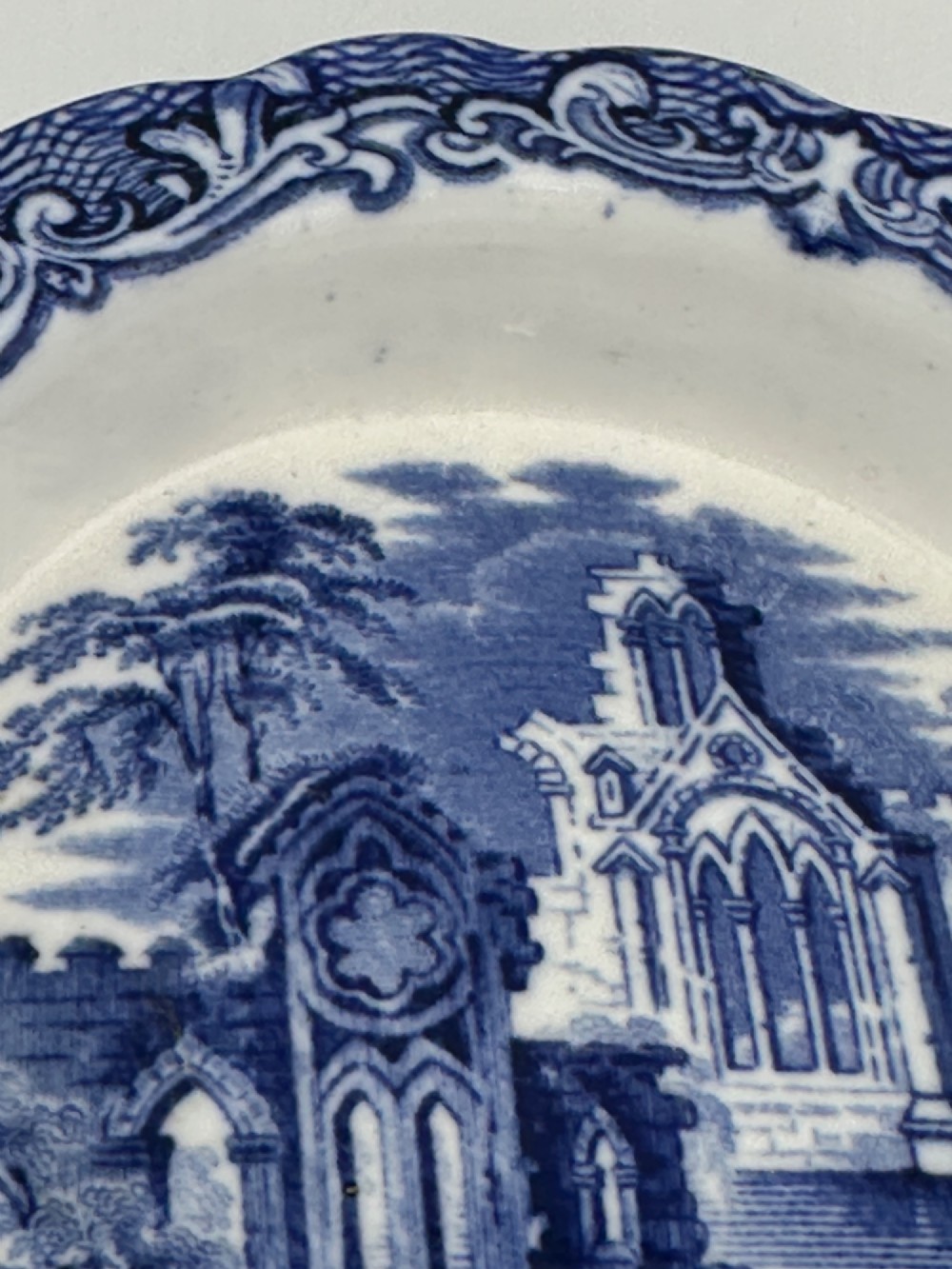 A circular blue and white transferware ceramic dish featuring a central printed depiction of abbey ruins framed by a decorative scrolled border. View VII