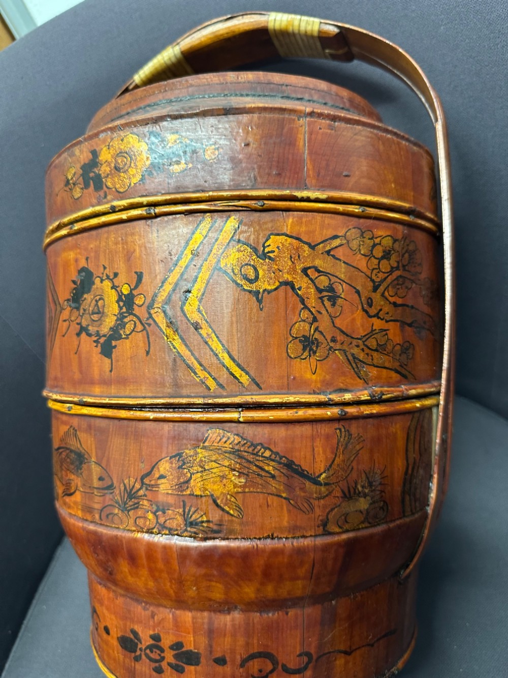 A three-tiered late 19th-century Chinese bamboo and wood food carrier with an arched handle, featuring hand-painted yellow flowers, birds, and goldfish against a warm brown lacquer finish. View XV