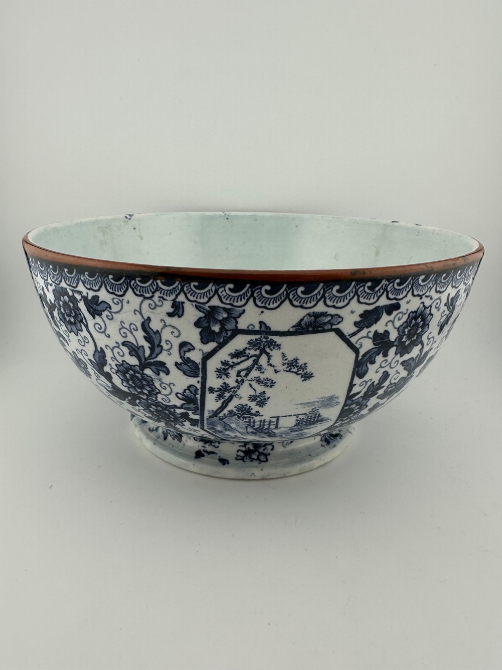 A deep circular footed earthenware bowl decorated with blue and white floral and landscape transferware patterns with a brown rim.