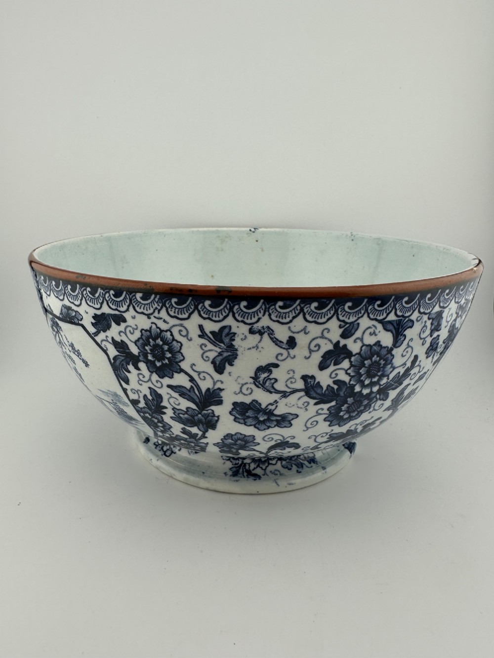 A deep circular footed earthenware bowl decorated with blue and white floral and landscape transferware patterns with a brown rim. View II