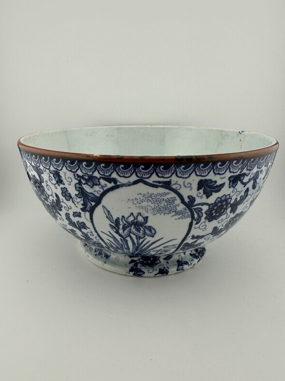 A deep circular footed earthenware bowl decorated with blue and white floral and landscape transferware patterns with a brown rim. View IV