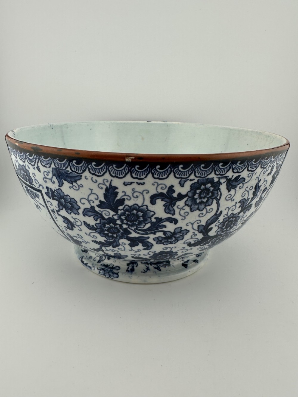 A deep circular footed earthenware bowl decorated with blue and white floral and landscape transferware patterns with a brown rim. View V