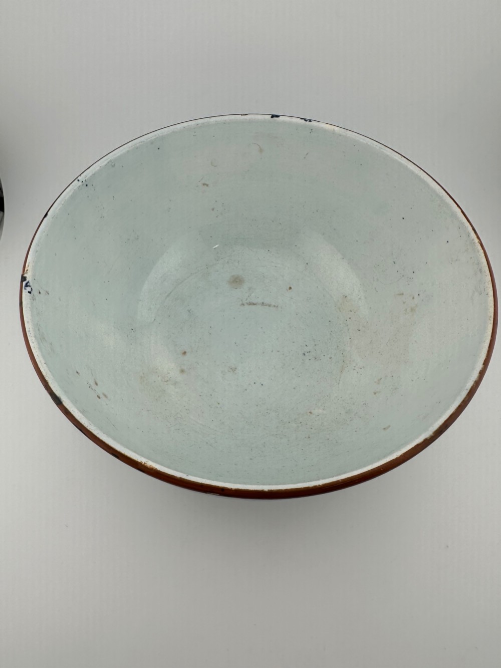 A deep circular footed earthenware bowl decorated with blue and white floral and landscape transferware patterns with a brown rim. View VI