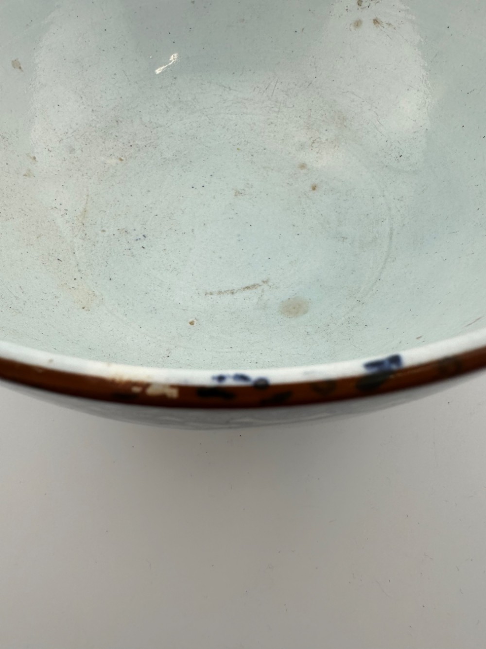 A deep circular footed earthenware bowl decorated with blue and white floral and landscape transferware patterns with a brown rim. View X