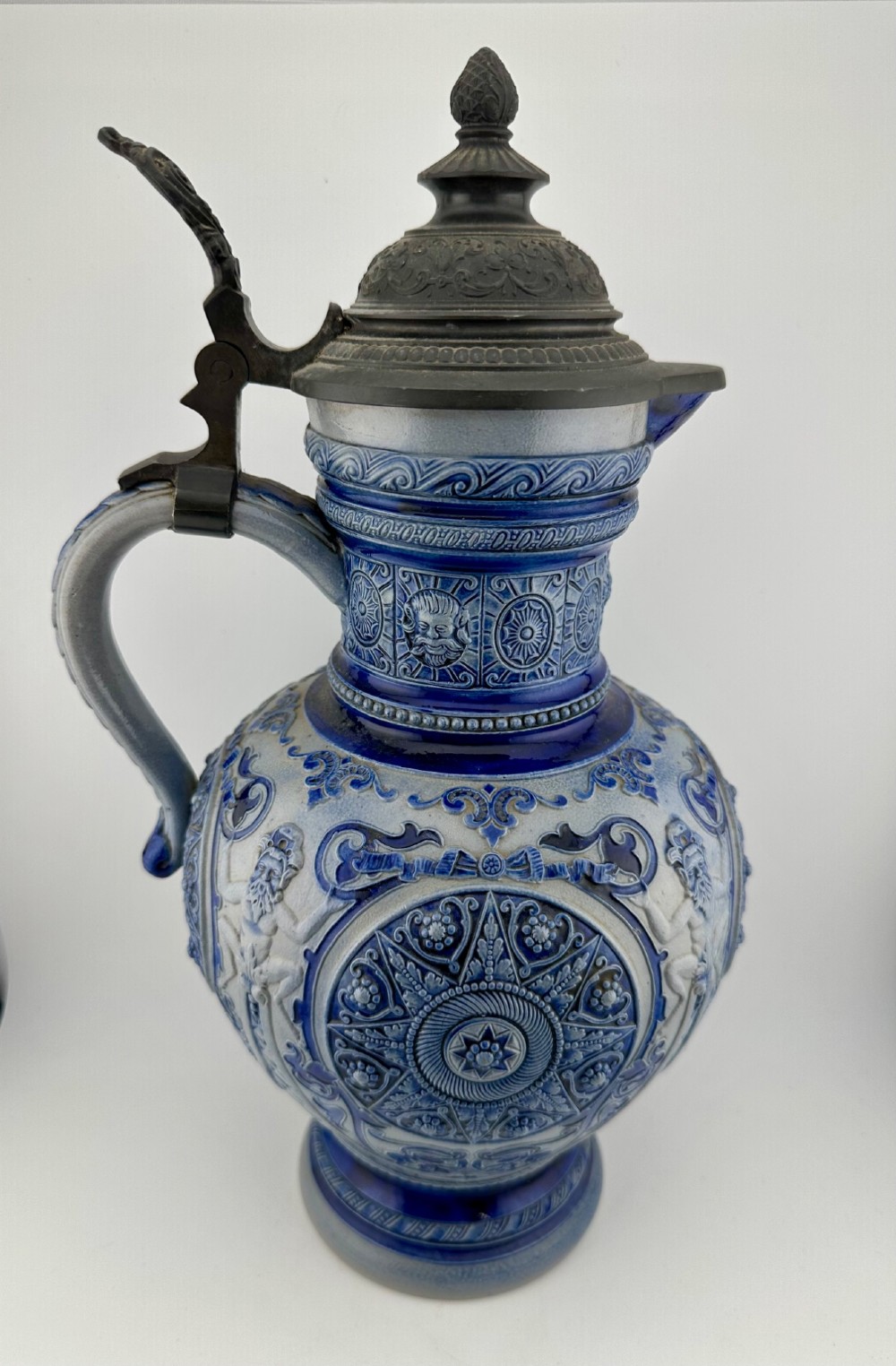 A blue and grey salt-glazed stoneware jug featuring relief-moulded geometric patterns, mask motifs, and a hinged pewter lid.