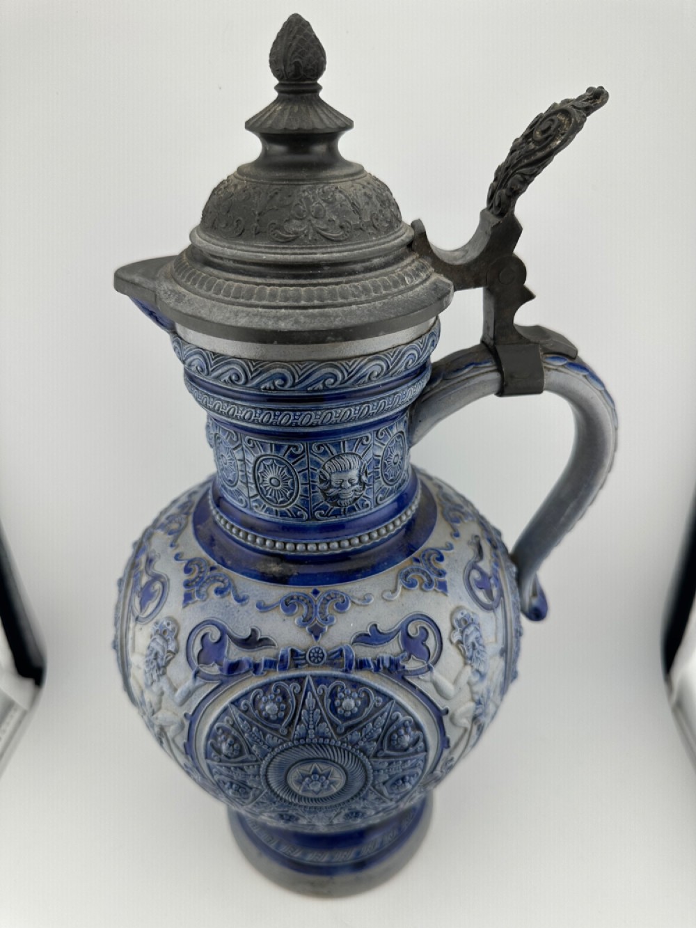 A blue and grey salt-glazed stoneware jug featuring relief-moulded geometric patterns, mask motifs, and a hinged pewter lid. View VI