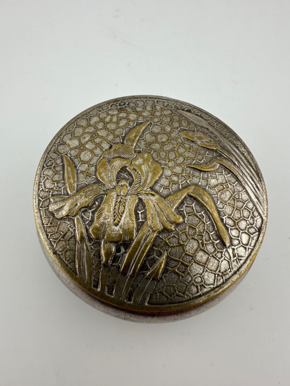 A circular antique Art Nouveau white metal compact box featuring a relief-carved iris floral design with brass highlights and a textured background.