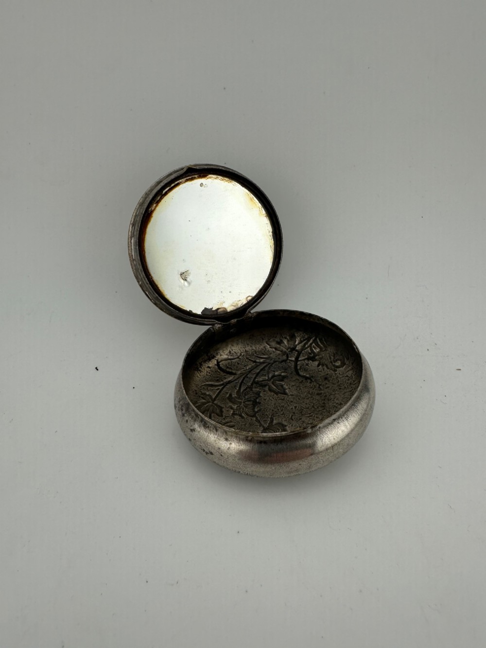 A circular antique Art Nouveau white metal compact box featuring a relief-carved iris floral design with brass highlights and a textured background. View II