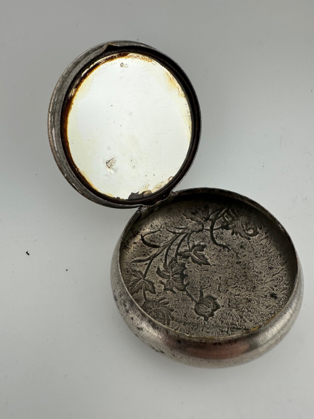 A circular antique Art Nouveau white metal compact box featuring a relief-carved iris floral design with brass highlights and a textured background. View III