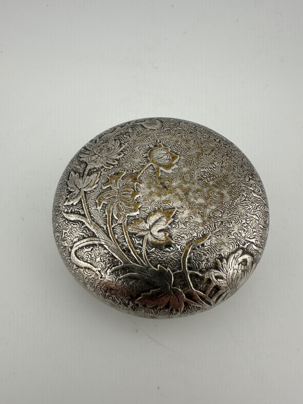 A circular antique Art Nouveau white metal compact box featuring a relief-carved iris floral design with brass highlights and a textured background. View V