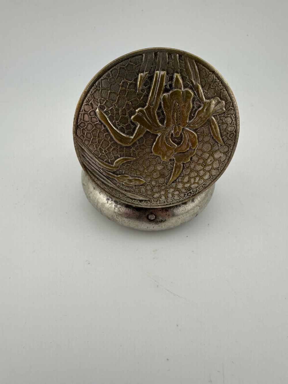 A circular antique Art Nouveau white metal compact box featuring a relief-carved iris floral design with brass highlights and a textured background. View VI