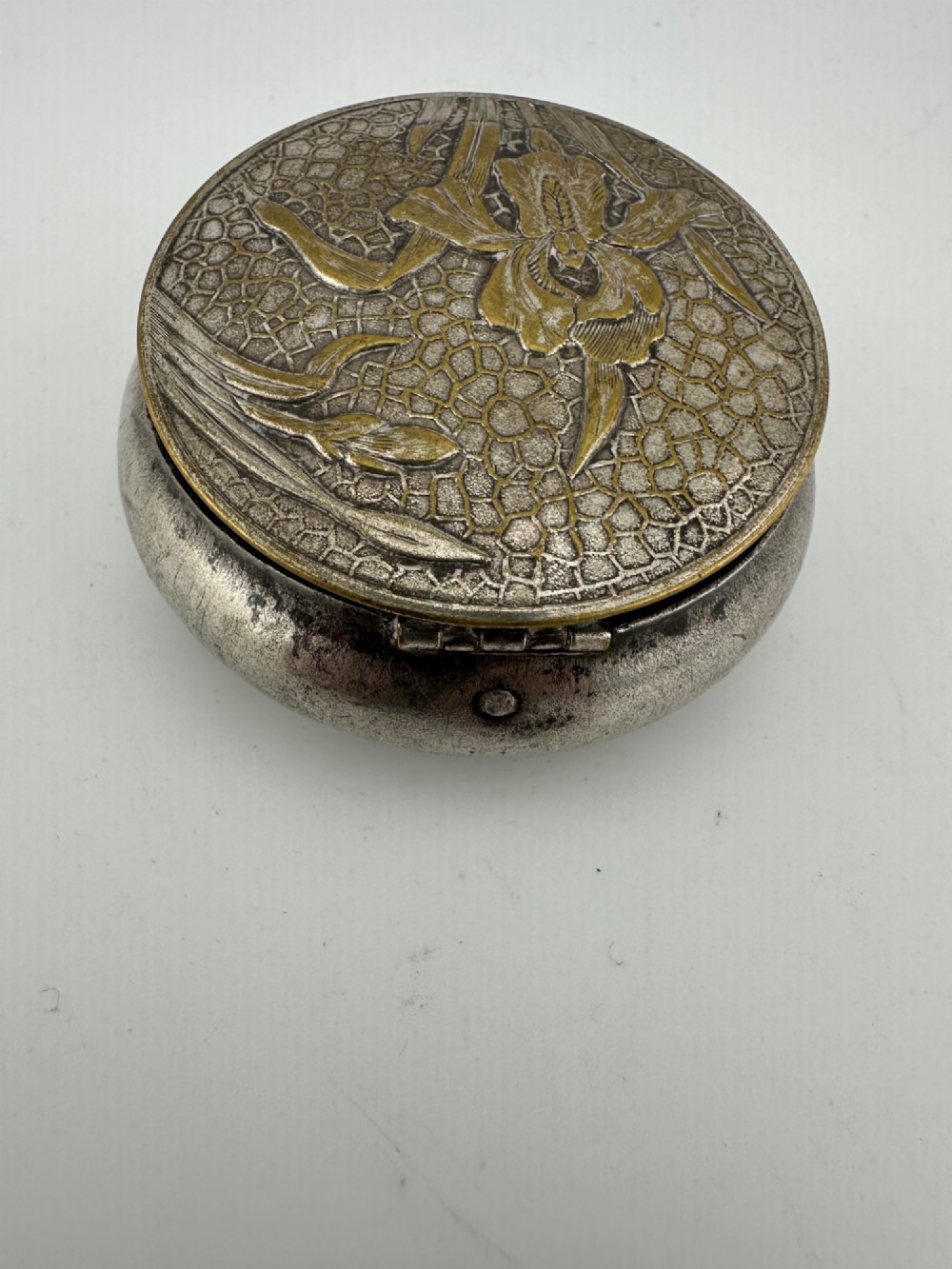 A circular antique Art Nouveau white metal compact box featuring a relief-carved iris floral design with brass highlights and a textured background. View VII