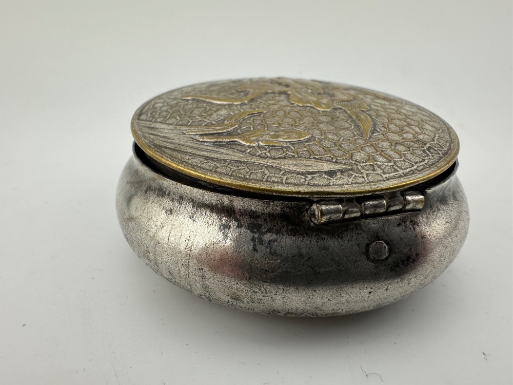 A circular antique Art Nouveau white metal compact box featuring a relief-carved iris floral design with brass highlights and a textured background. View VIII