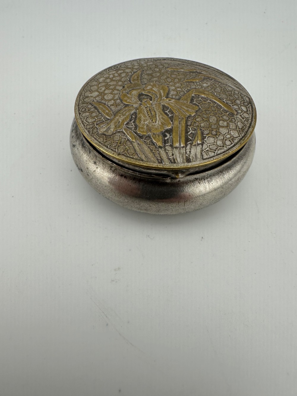 A circular antique Art Nouveau white metal compact box featuring a relief-carved iris floral design with brass highlights and a textured background. View X