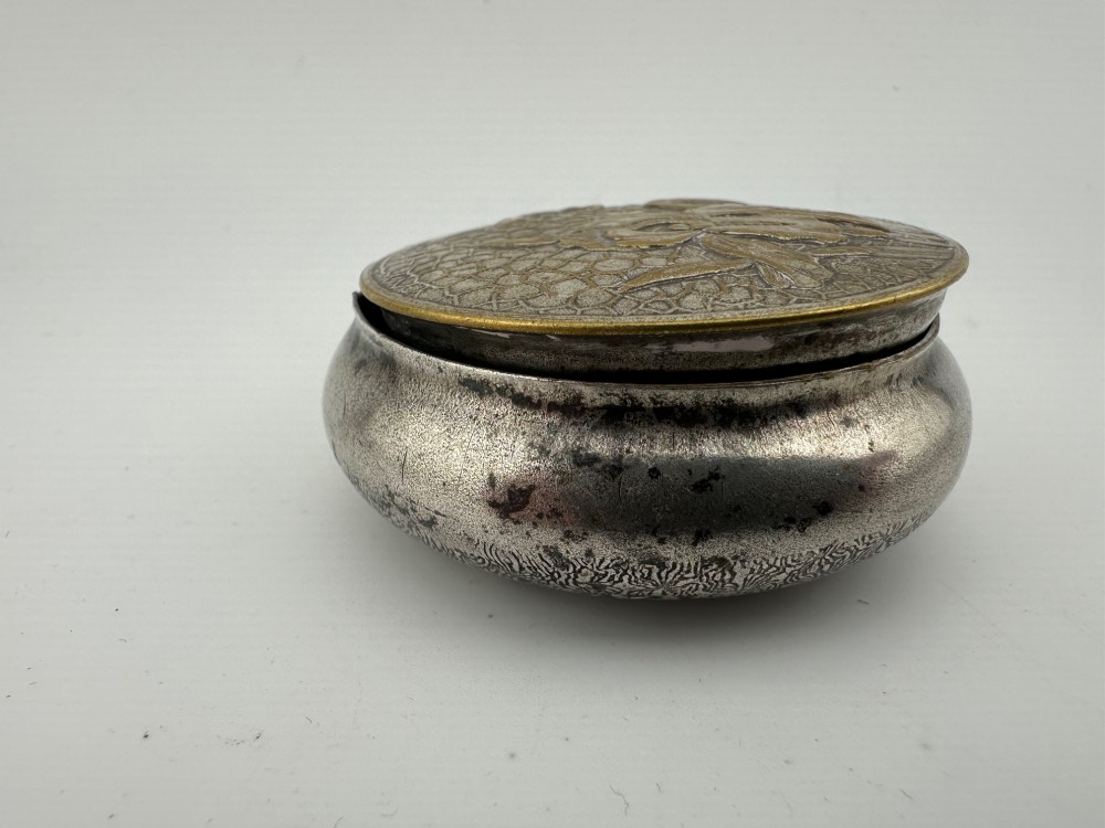 A circular antique Art Nouveau white metal compact box featuring a relief-carved iris floral design with brass highlights and a textured background. View XI