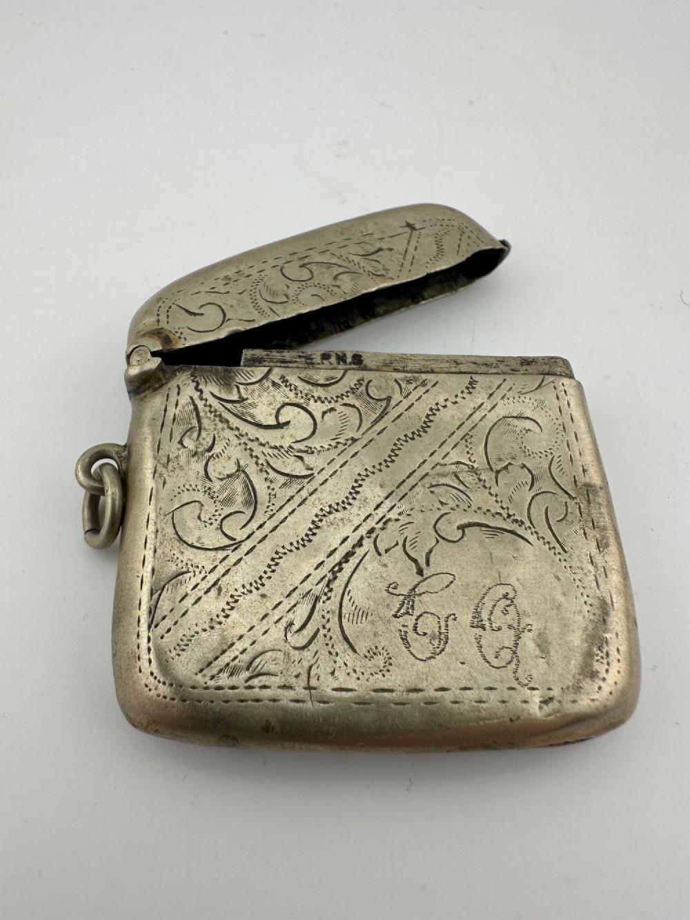 An antique rectangular silver-plated vesta case featuring hand-engraved scrollwork, an open hinged lid, and a side suspension ring.