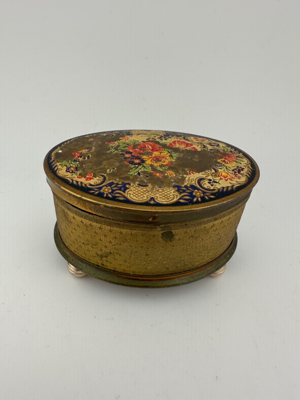 An oval vintage metal trinket box featuring a printed floral motif on the lid, star-patterned side panels, small rounded feet, and a velvet lining.
