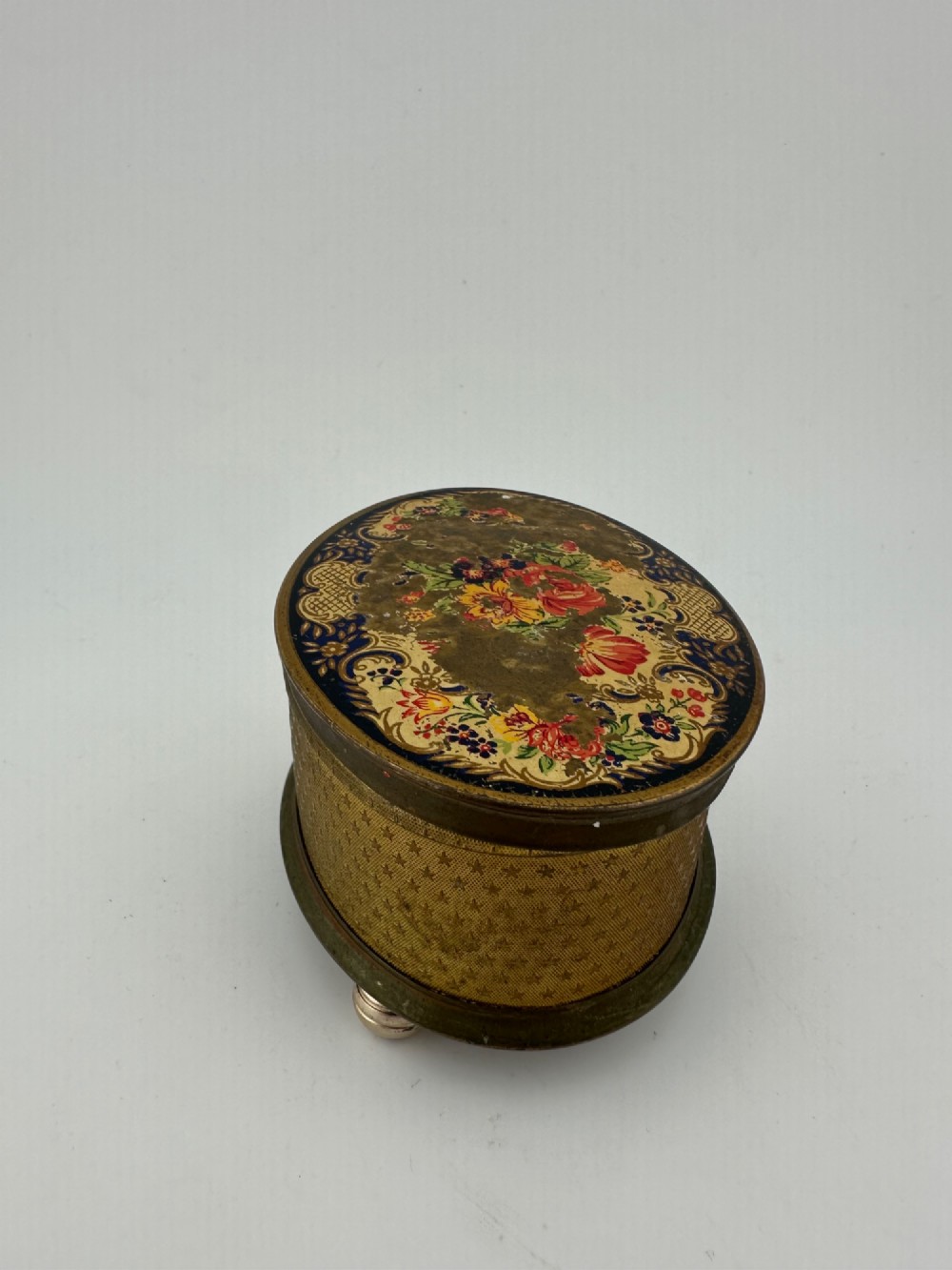 An oval vintage metal trinket box featuring a printed floral motif on the lid, star-patterned side panels, small rounded feet, and a velvet lining. View V