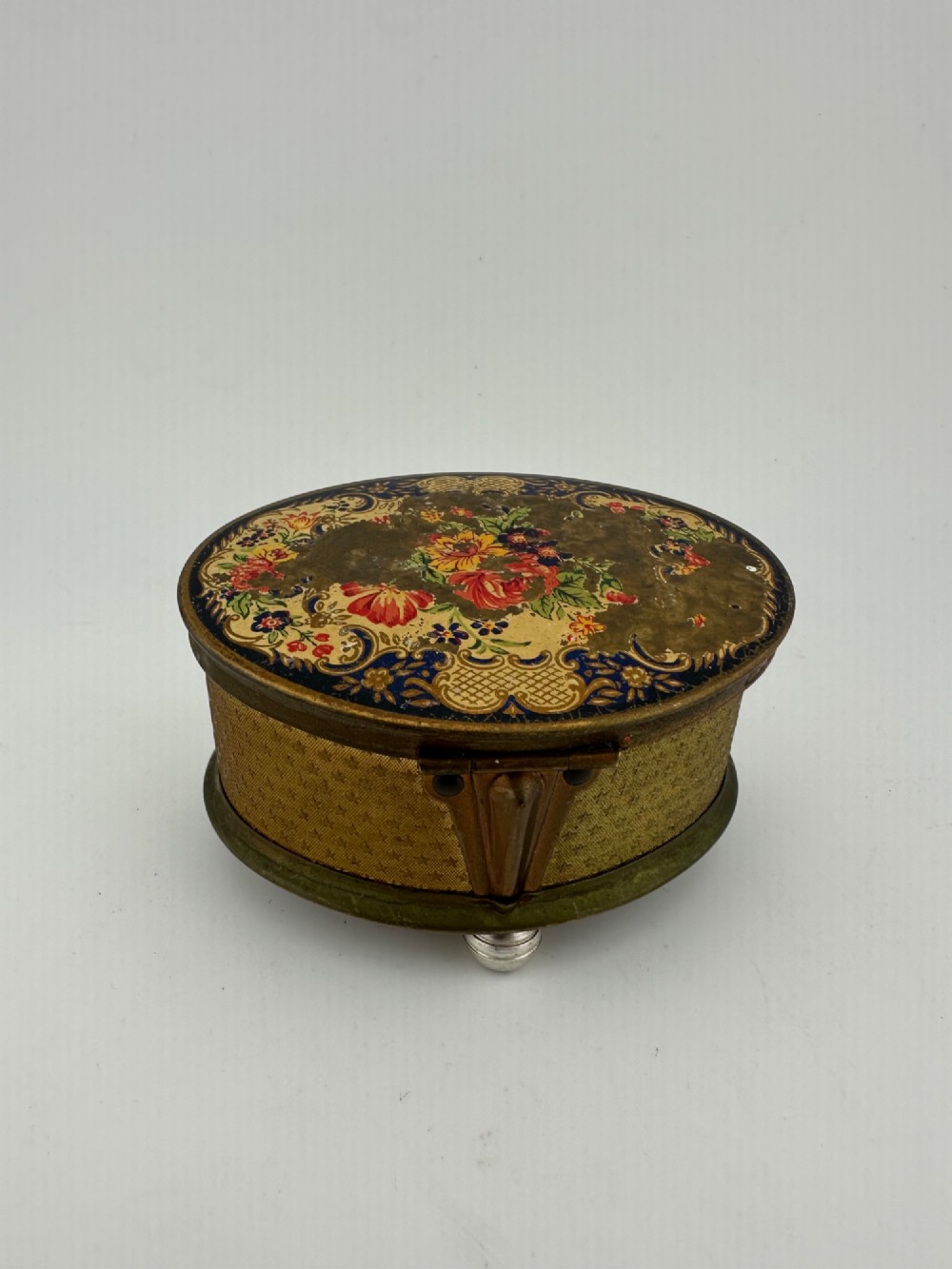 An oval vintage metal trinket box featuring a printed floral motif on the lid, star-patterned side panels, small rounded feet, and a velvet lining. View VI