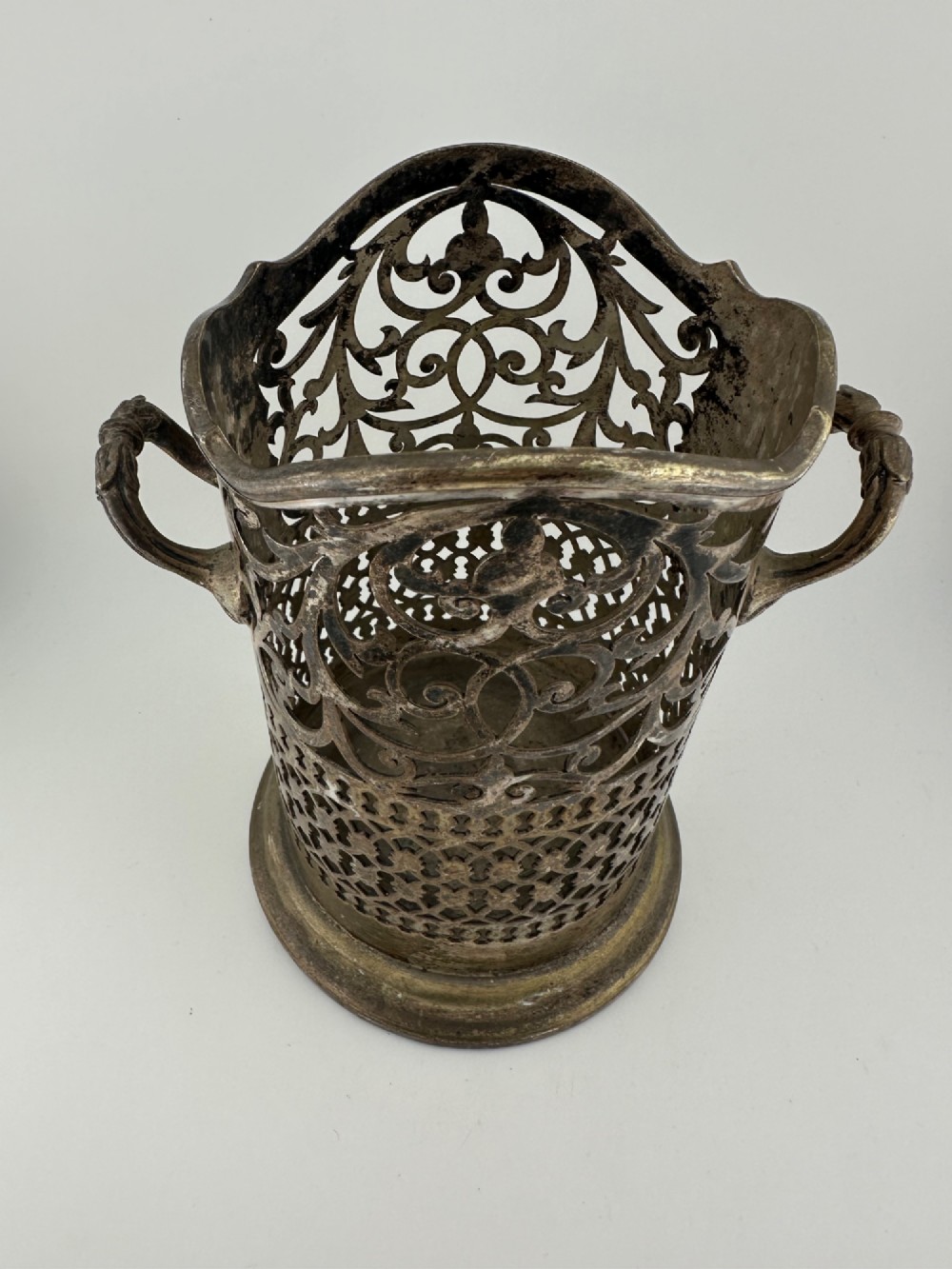 An antique silver-plated cylindrical wine bottle holder featuring pierced scroll fretwork, two side handles, and a flared circular base. View IV