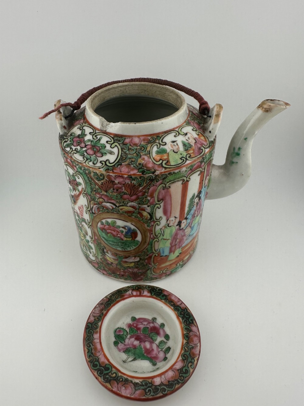 A Chinese Famille Rose cylindrical ceramic teapot with hand-painted court figures set beside its fitted woven wicker carrying case. View X