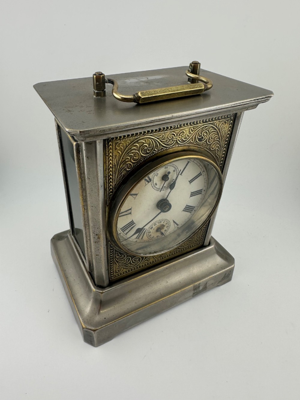 An Edwardian carriage clock with a silvered metal casing, brass carrying handle, and engraved brass mask around a white enamel dial. View II