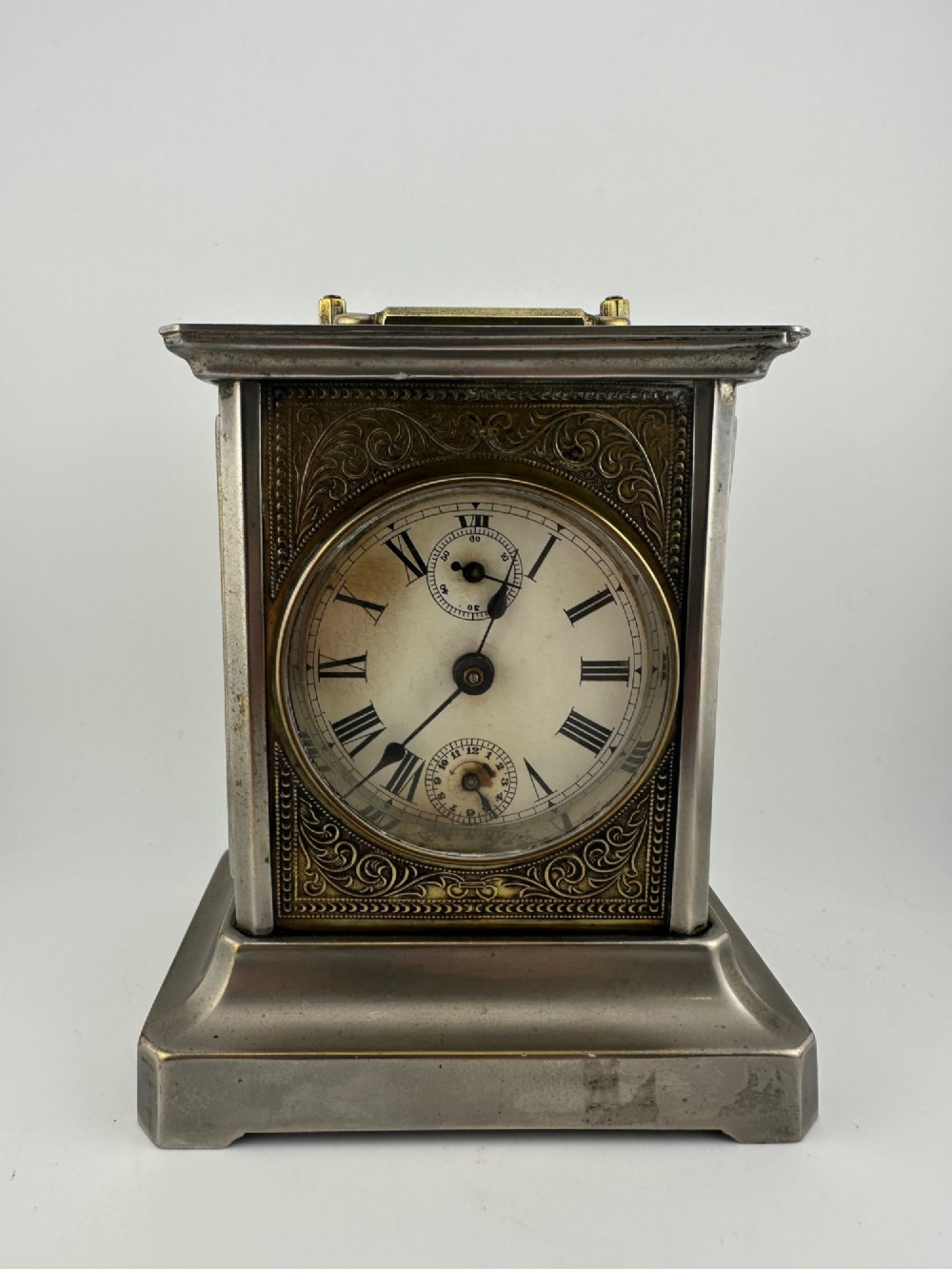 An Edwardian carriage clock with a silvered metal casing, brass carrying handle, and engraved brass mask around a white enamel dial. View III