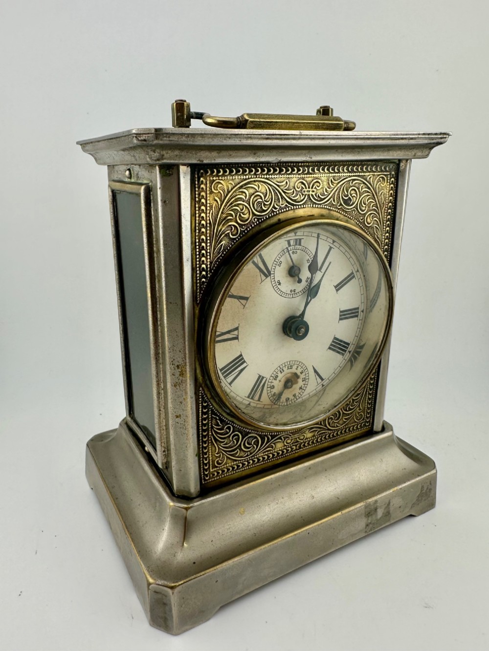 An Edwardian carriage clock with a silvered metal casing, brass carrying handle, and engraved brass mask around a white enamel dial.