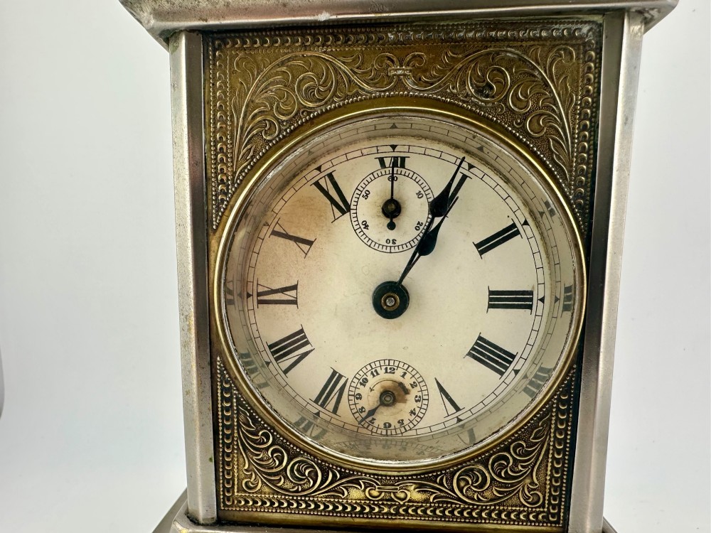 An Edwardian carriage clock with a silvered metal casing, brass carrying handle, and engraved brass mask around a white enamel dial. View XIV
