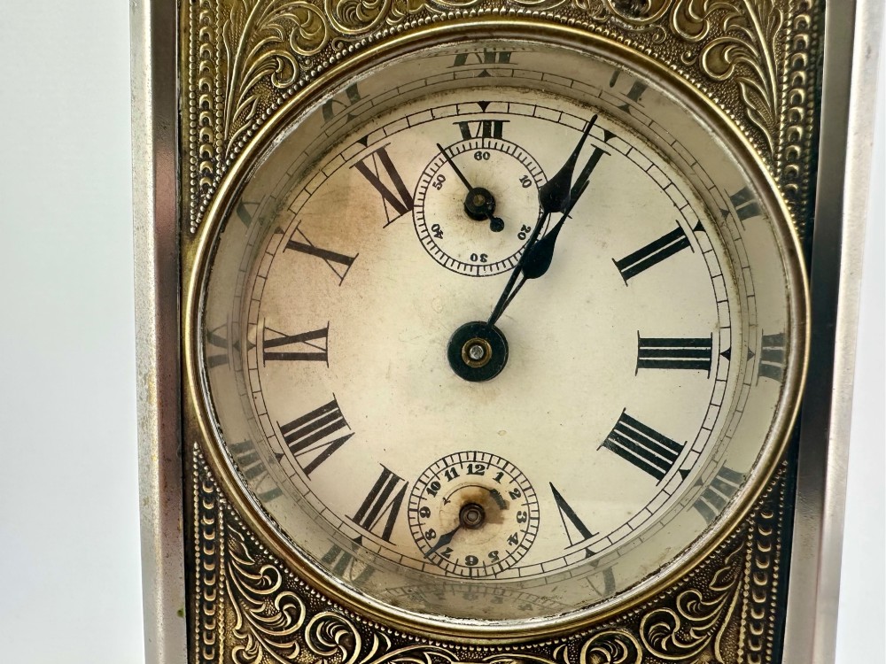An Edwardian carriage clock with a silvered metal casing, brass carrying handle, and engraved brass mask around a white enamel dial. View XV