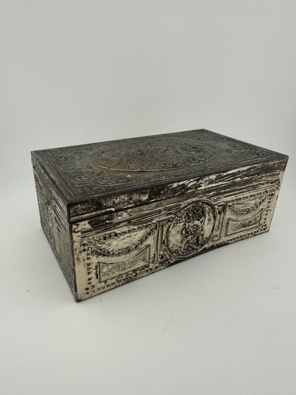 A rectangular silver-grey metal box featuring embossed neoclassical cherub medallions and relief swags across its hinged lid and sides.