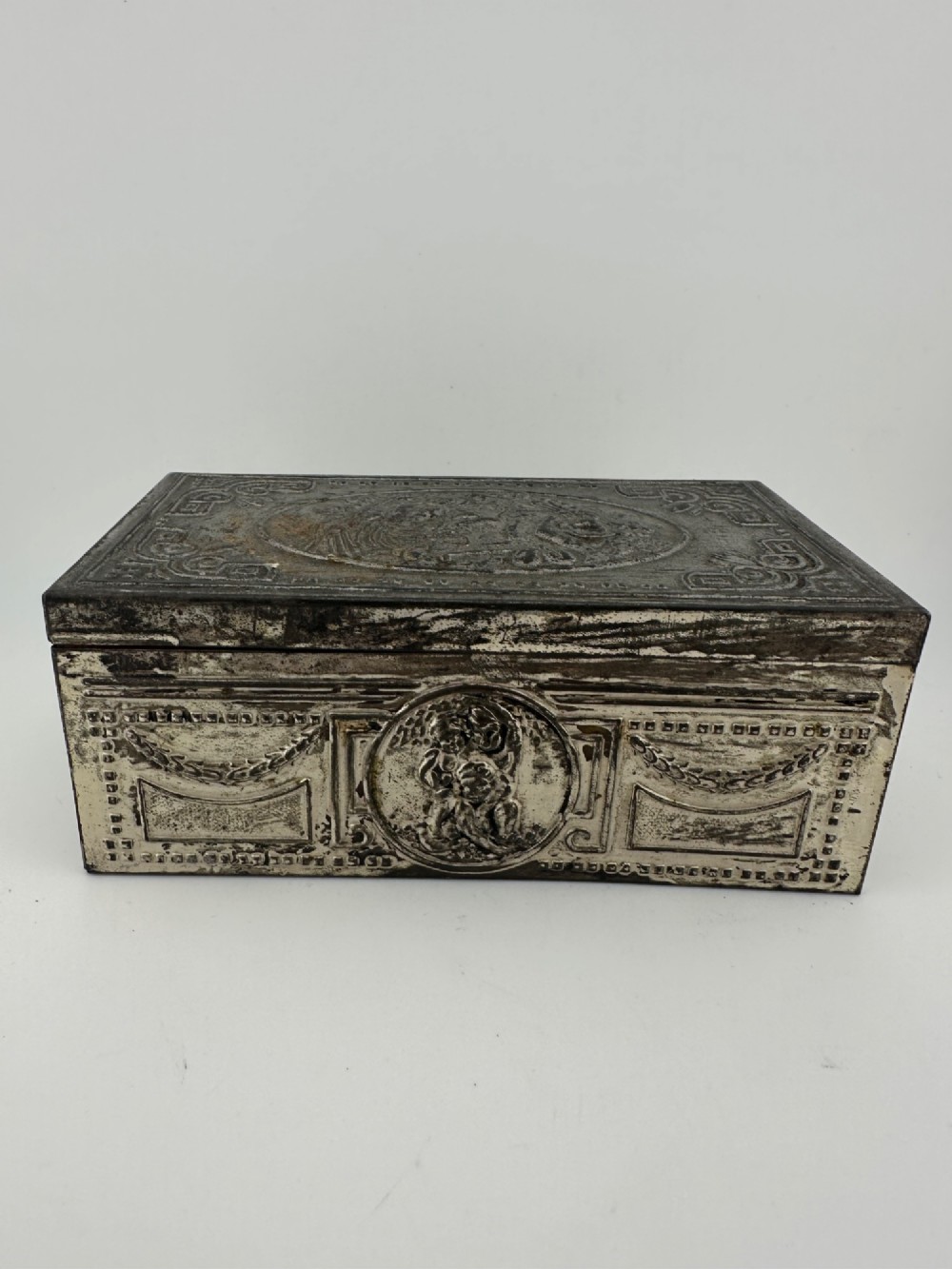 A rectangular silver-grey metal box featuring embossed neoclassical cherub medallions and relief swags across its hinged lid and sides. View II