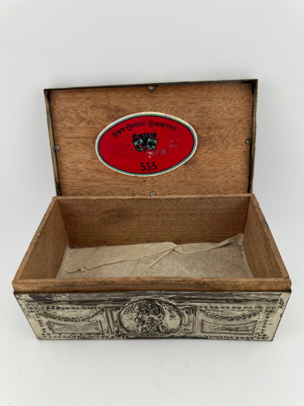 A rectangular silver-grey metal box featuring embossed neoclassical cherub medallions and relief swags across its hinged lid and sides. View III