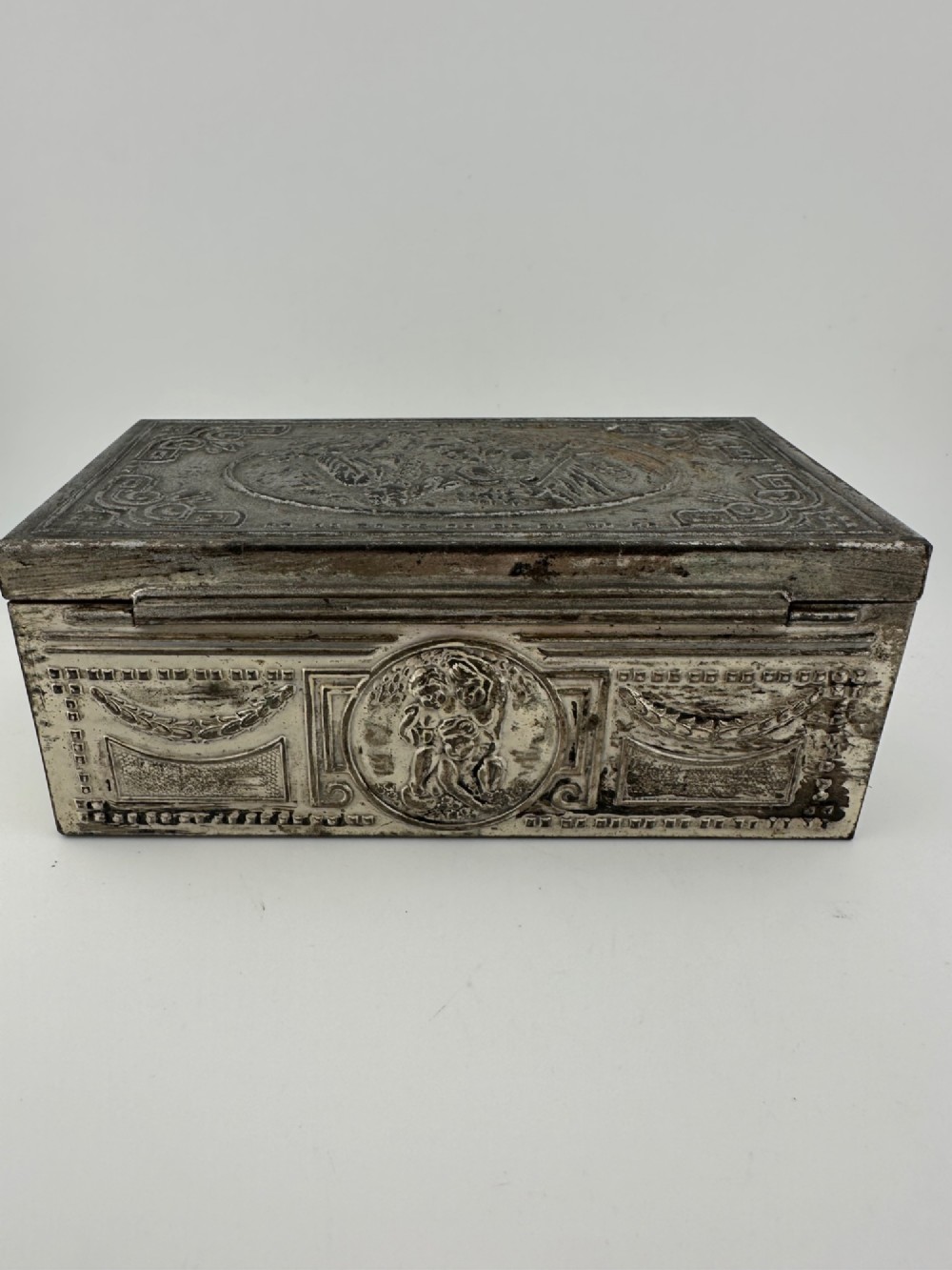 A rectangular silver-grey metal box featuring embossed neoclassical cherub medallions and relief swags across its hinged lid and sides. View IV