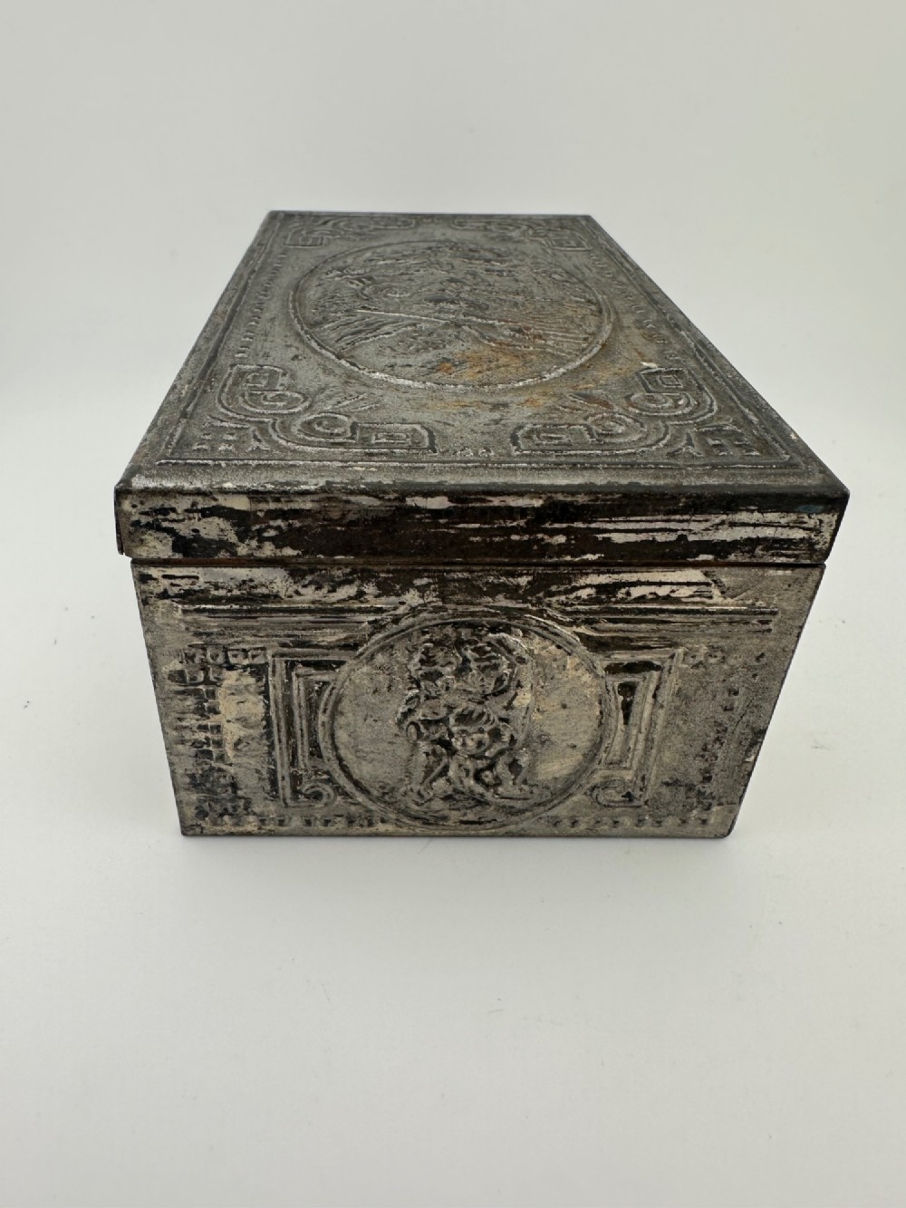 A rectangular silver-grey metal box featuring embossed neoclassical cherub medallions and relief swags across its hinged lid and sides. View VI