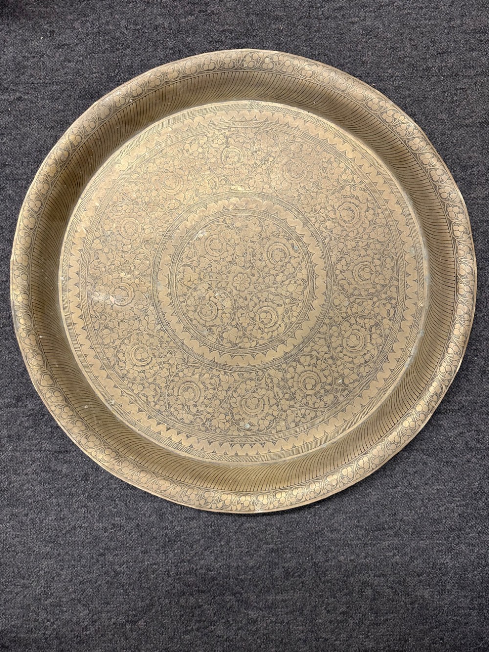 A large circular brass tray featuring intricate hand-engraved concentric floral and geometric patterns with a shallow rolled rim.