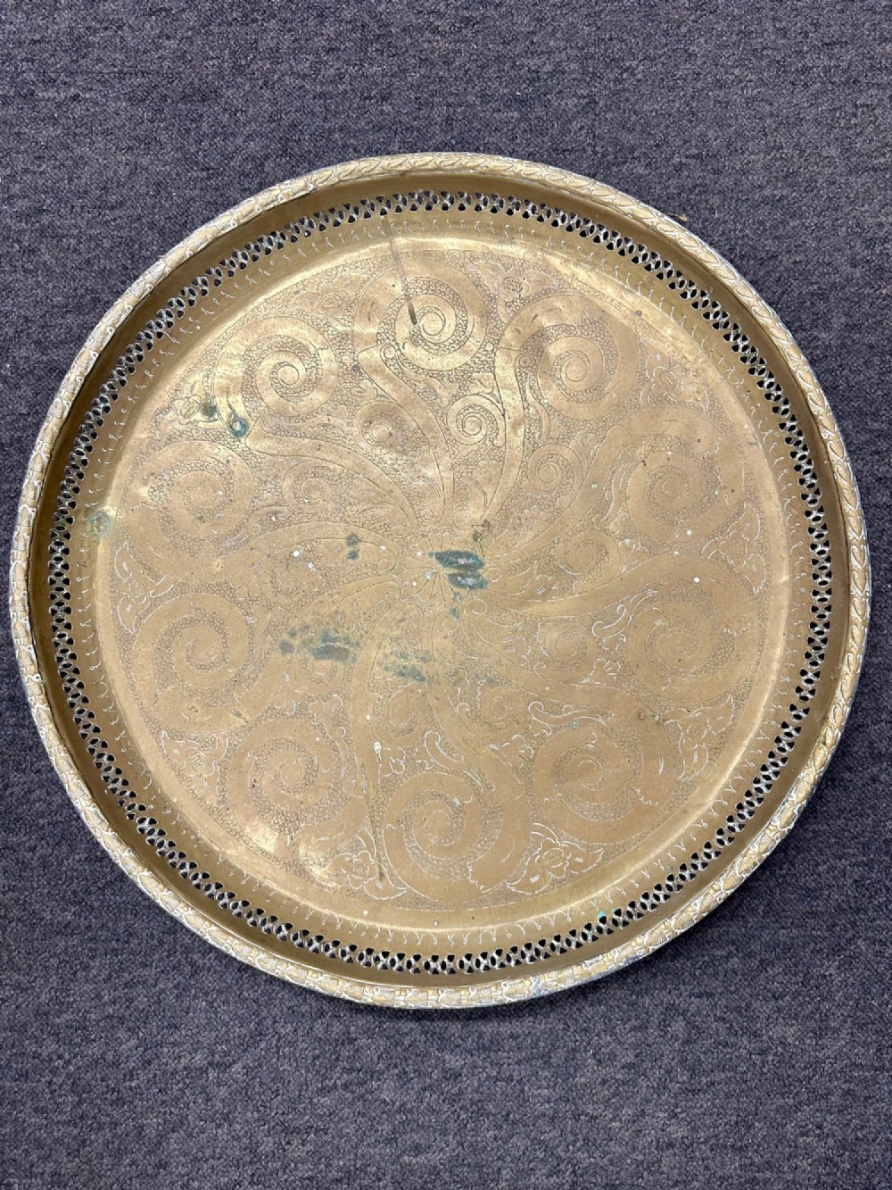 A circular antique brass tray featuring an intricately engraved swirling floral pattern and a pierced gallery rim.