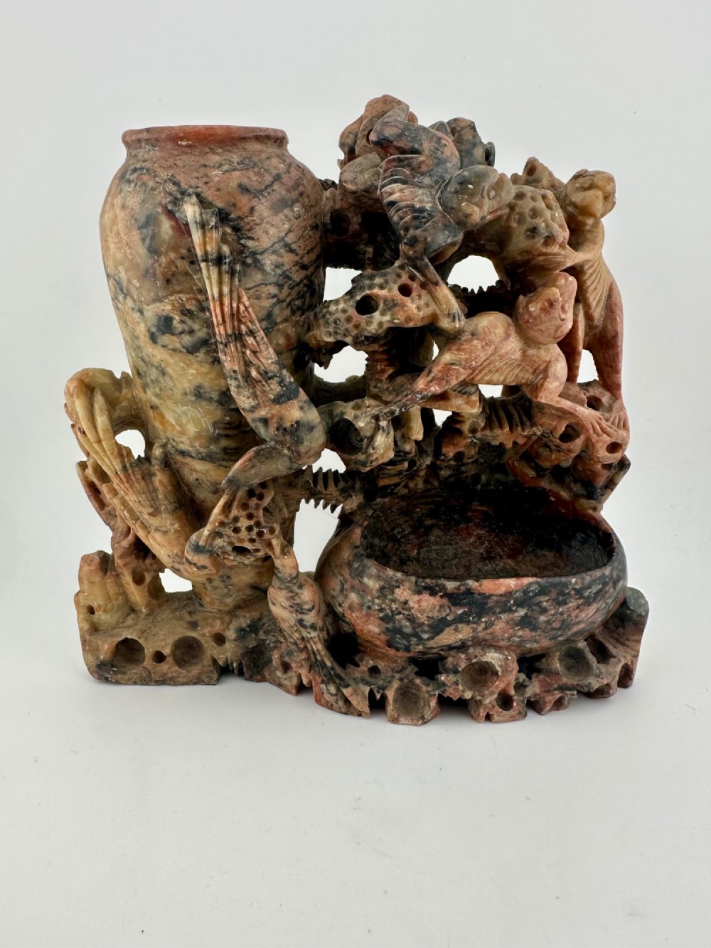 A Qing dynasty Chinese soapstone carving depicting a small vase, a shallow brush washer basin, and carved birds among naturalistic floral motifs in mottled red, grey, and cream stone.