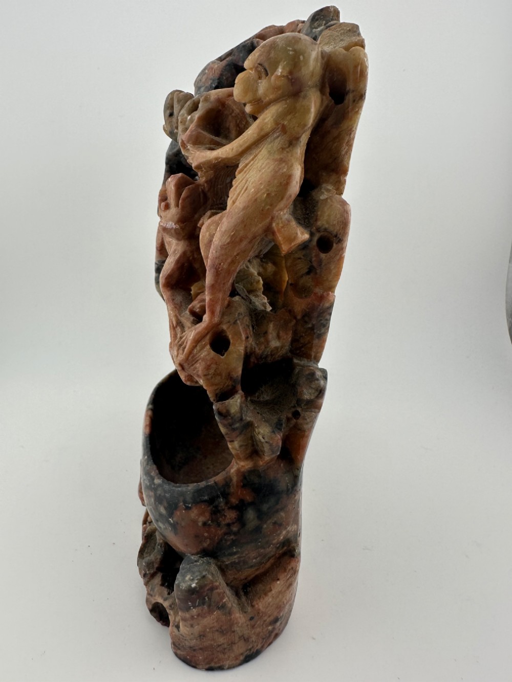 A Qing dynasty Chinese soapstone carving depicting a small vase, a shallow brush washer basin, and carved birds among naturalistic floral motifs in mottled red, grey, and cream stone. View X