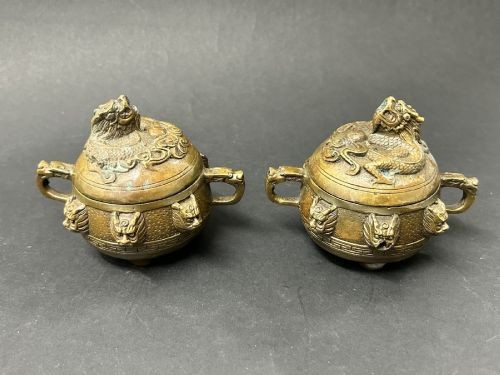 A matching pair of rounded bronze censers featuring relief dragon heads around the body, twin side handles, and sculpted dragon lid finials.