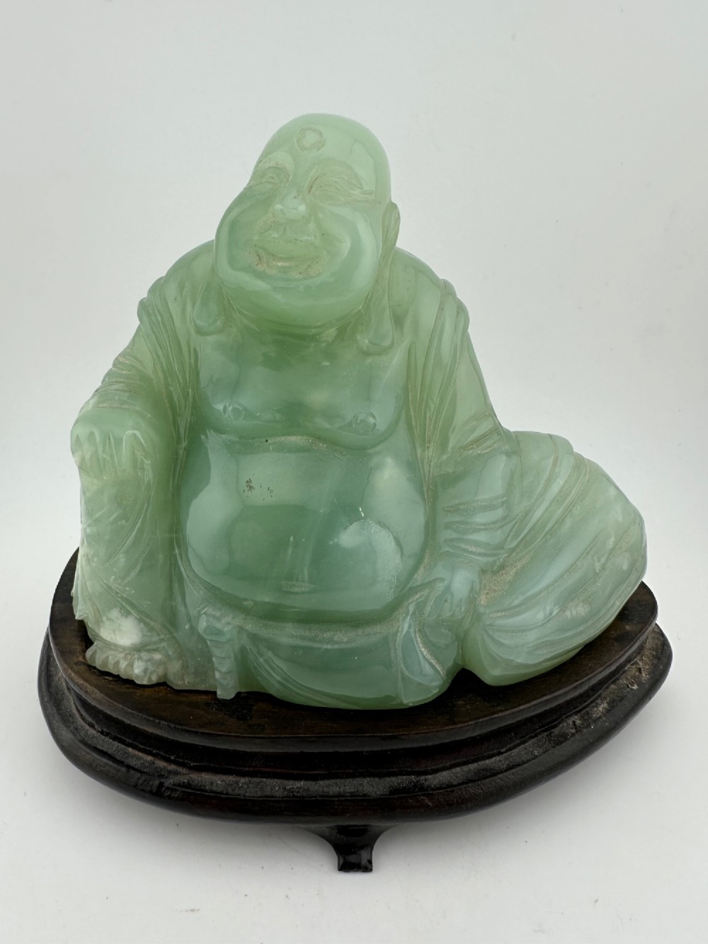 A hand-carved light green jadeite sculpture depicting a seated Laughing Buddha resting on a dark wooden fitted base.