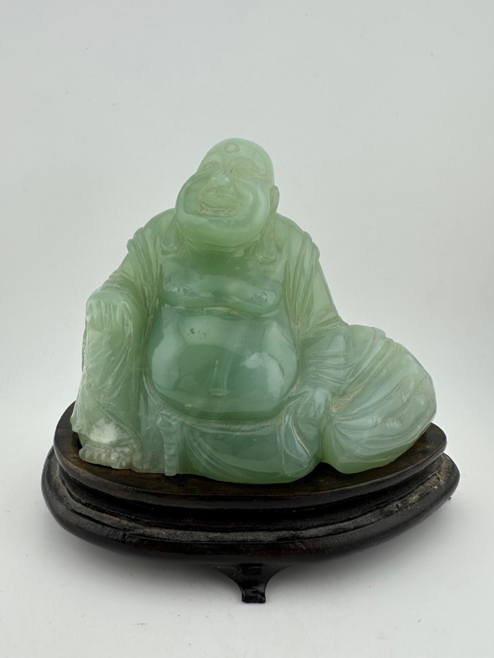 A hand-carved light green jadeite sculpture depicting a seated Laughing Buddha resting on a dark wooden fitted base. View II