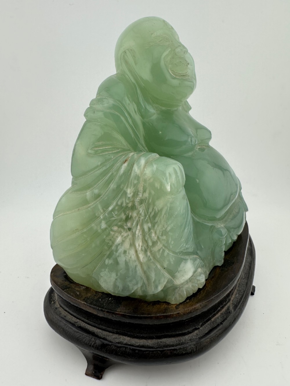 A hand-carved light green jadeite sculpture depicting a seated Laughing Buddha resting on a dark wooden fitted base. View IV